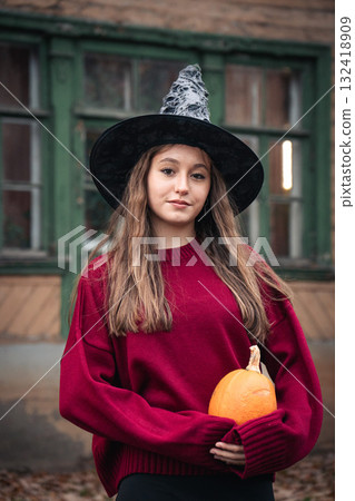 Young woman in a witch hat and burgundy sweater holds a pumpkin in front of an old wooden house with green window frames Young woman in a witch hat and burgundy sweater holds a pumpkin in front of an old wooden house with green window frames 132418909