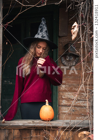 Young woman in a witch hat and red sweater stands near a rustic wooden house with a pumpkin on the railing, surrounded by dry vines 132418911