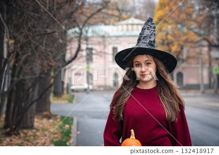 Young woman in a witch hat and burgundy sweater holding a pumpkin walks along an autumn city street. Halloween portrait Young woman in a witch hat and burgundy sweater holding a pumpkin walks along an autumn city street. Halloween portrait 132418917