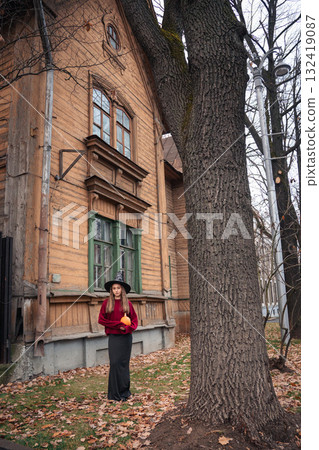 Woman in a witch hat and burgundy sweater holds a pumpkin in front of an old wooden house with green windows Woman in a witch hat and burgundy sweater holds a pumpkin in front of an old wooden house with green windows 132419087