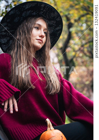 Young woman in a burgundy sweater and black hat sits on a park bench beside a small pumpkin, surrounded by autumn foliage Young woman in a burgundy sweater and black hat sits on a park bench beside a small pumpkin, surrounded by autumn foliage 132419091