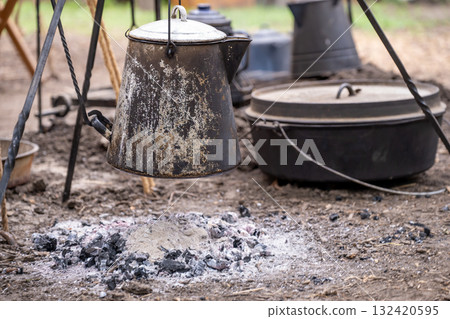 Cooking kettle hangs over campfire ash beside black cooking pots during outdoor gathering Cooking kettle hangs over campfire ash beside black cooking pots during outdoor gathering 132420595