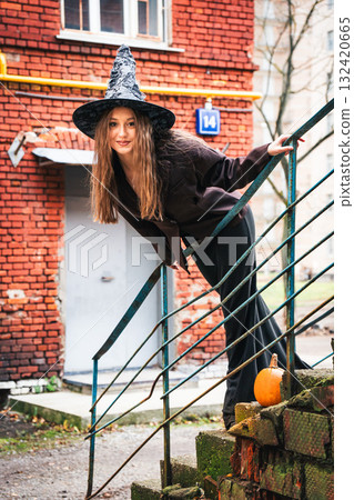 Smiling teen girl in a witch hat leans playfully on old stairs with a pumpkin nearby 132420665