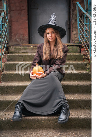 Teen girl in a witch hat and long dark skirt sits on old stone steps holding a pumpkin Teen girl in a witch hat and long dark skirt sits on old stone steps holding a pumpkin 132420669
