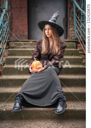 Teen girl in a witch hat and long dark skirt sits on old stone steps holding a pumpkin 132420670