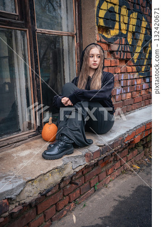 Young woman in black clothes and boots sits on a brick windowsill next to a pumpkin 132420671