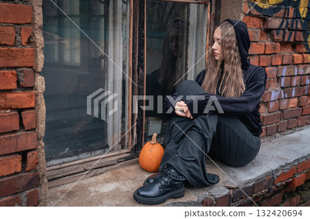 Young woman in black clothes and boots sits on a brick windowsill next to a pumpkin 132420694