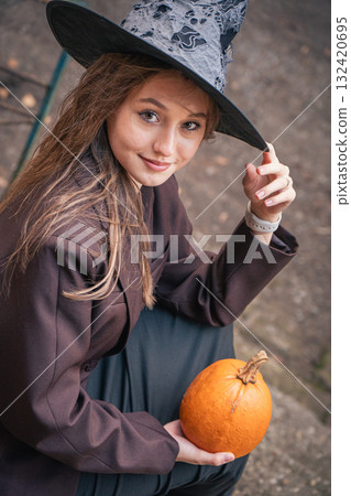 Smiling teen girl in a witch hat and brown blazer holds a small pumpkin outdoors 132420695