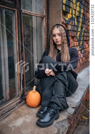 Young woman in black clothes and boots sits on a brick windowsill next to a pumpkin 132420732