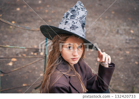 Teen girl in a witch hat looks into the camera while adjusting the brim, creating a confident Halloween portrait 132420733