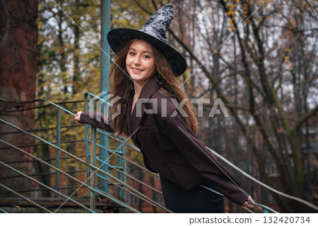 Smiling teen girl in a witch hat leans playfully on a rusty staircase outdoors. Cheerful Halloween portrait 132420734