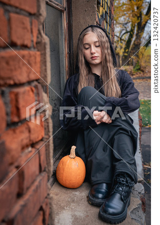 A yuong woman in dark clothes with hood and boots sits by an old brick wall next to a small pumpkin 132420737