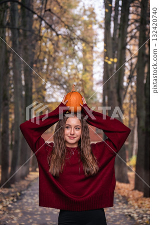 Teen girl in a red sweater holding a small pumpkin on her head in a park with autumn trees. Great for Halloween Teen girl in a red sweater holding a small pumpkin on her head in a park with autumn trees. Great for Halloween 132420740