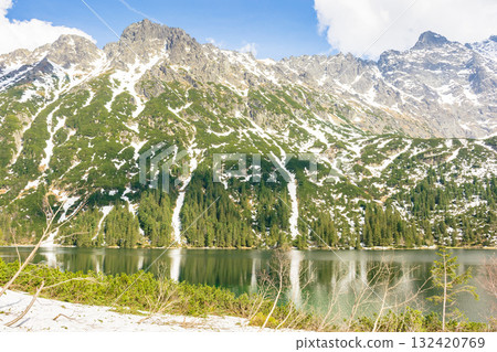 alpine lake in spring on a sunny day. travel landscape of rysy ridge in high tatra mountains of poland. snow among rocks and spruce forest on the hillside reflection in water. blue sky with clouds 132420769