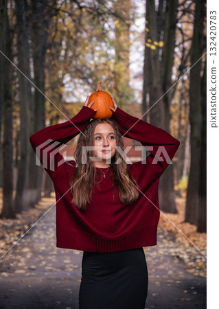 Teen girl in a red sweater holding a small pumpkin on her head in a park with autumn trees. Great for Halloween 132420783