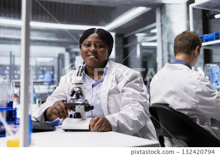 Portrait of black woman posing in laboratory at desk with microscope, examining specimen for a research study. Scientist in white coat emphasizing cellular discovery in bioscience. Portrait of black woman posing in laboratory at desk with microscope, examining specimen for a research study. Scientist in white coat emphasizing cellular discovery in bioscience. 132420794