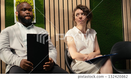 Black male job seeker waiting to be called after a person in front of him, sitting in queue on the hallway with his registration files and CV in hand. Calling in candidates. 132420862