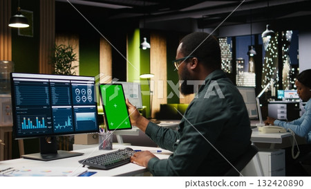 Green screen on tablet next to young black businessman reviewing data, working on analytics report writing and finding solutions for global expansion and business management tasks. Green screen on tablet next to young black businessman reviewing data, working on analytics report writing and finding solutions for global expansion and business management tasks. 132420890