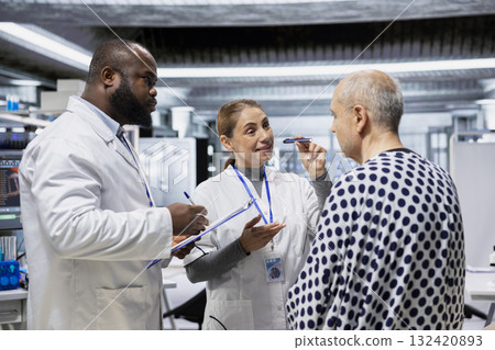 Diverse team of doctors examining a senior man for a new drug testing study, monitoring symptoms or potential allergic reactions. Looking for side effects under observation in lab. 132420893