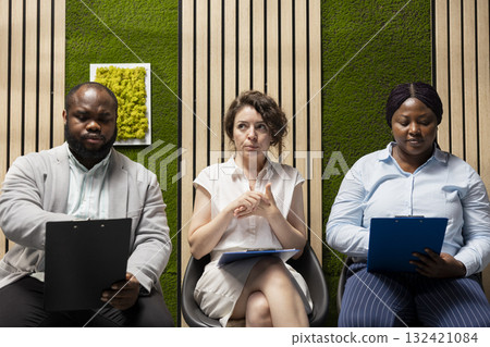Anxious diverse job seekers awaiting job interview discussion with manager, showcasing anticipation in office hallway. People preparing with information for the recruitment process. Anxious diverse job seekers awaiting job interview discussion with manager, showcasing anticipation in office hallway. People preparing with information for the recruitment process. 132421084