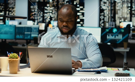 Black office worker reclining in a chair and relaxing at midnight, daydreaming during a work break. Fulfilled and self assured, reflecting on career success and corporate achievements. 132421109