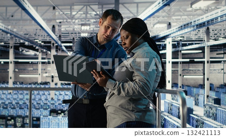 Server room coworkers scan infrastructure for security issues using laptop. Data center system administrators using notebook to organize file backups and plan recovery steps, camera B 132421113