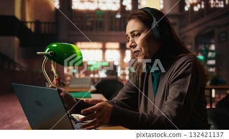 Female student participates in online class via laptop in a campus library, engaged in video conference lecture while researching for a thesis and studying for academic qualifications. Female student participates in online class via laptop in a campus library, engaged in video conference lecture while researching for a thesis and studying for academic qualifications. 132421137