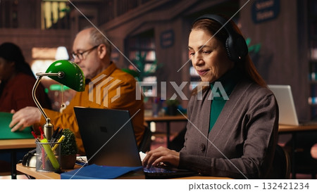 Mature student listening to school webinar on laptop at a library, takes notes and does research during online lecture. Studying remotely with an expert to obtain her diploma. 132421234