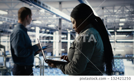 Data center admin investigates AI configurations using laptop, making adjustments to ensure optimal function. African american woman in server hub checks deep learning errors reports, camera B Data center admin investigates AI configurations using laptop, making adjustments to ensure optimal function. African american woman in server hub checks deep learning errors reports, camera B 132421250