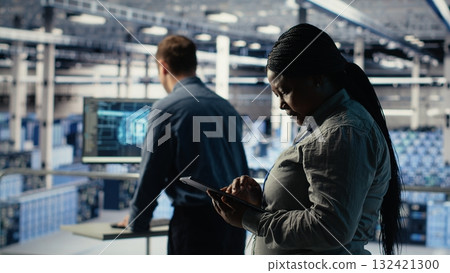 Software developers monitor server hub activity using tablet. Data center employees doing brainstorming, reviewing equipment tests results on devices to ensure systems are working correctly, camera B 132421300