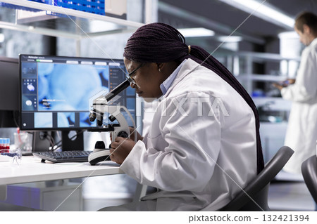 Black female researcher studies molecular biology samples in a lab, using professional microscope. Healthcare innovation and biochemistry experiments represent scientific method and discovery. 132421394