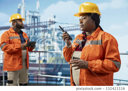Offshore platform professional using hand portable radio device to coordinate workflow during operations. African american drilling rig worker speaking in walkie talkie, doing emergency procedures 132421516