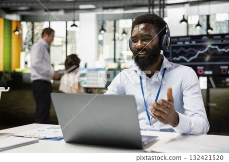 Business consultant wearing headphones engaging in video conference on laptop, explaining company strategies to remote clients. Black male manager in office, leading online meeting. Business consultant wearing headphones engaging in video conference on laptop, explaining company strategies to remote clients. Black male manager in office, leading online meeting. 132421520