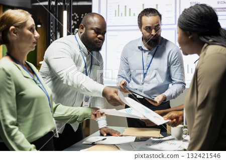 Multiethnic work team exchanging documents and using data visualization to forecast business goals in a quiet corporate strategy room. Staff members collaborating late at night. 132421546