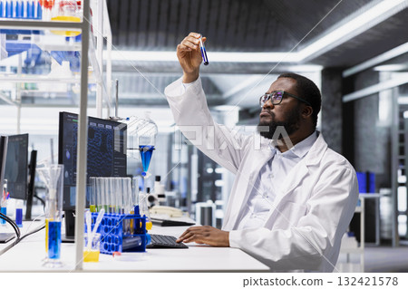 Molecular biologist using laboratory glassware filled with blue indicator solution at workstation in research facility. African american man picks sample tube with chemical fluid used for experiments 132421578