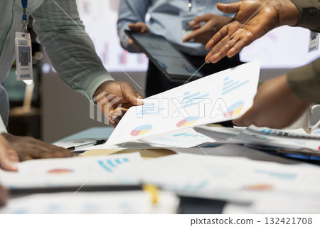 Close up of multiethnic workers shares insight on documents and reports during advisory board session late at night. Business associates reviewing diagrams for upcoming presentation. 132421708