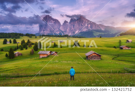 Hiker standing in a green meadow with wooden huts in mountains Hiker standing in a green meadow with wooden huts in mountains 132421752