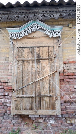 Closed old wooden shutters with rustic carved designs, with intricate carvings at the top. Shutters are antique and installed against a brick wall background. Closed old wooden shutters with rustic carved designs, with intricate carvings at the top. Shutters are antique and installed against a brick wall background. 132422095