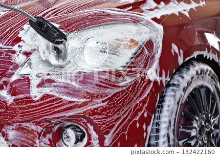 Front head light of red car washed in self serve carwash, brush leaving white soap strokes on glass, closeup detail 132422160