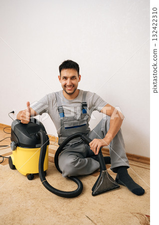 Friendly cleaning service employee in overall showing thumbs up gesture sitting on floor with washing vacuum cleaner, ready for thorough floor cleaning and sanitizing service, looking at camera. Friendly cleaning service employee in overall showing thumbs up gesture sitting on floor with washing vacuum cleaner, ready for thorough floor cleaning and sanitizing service, looking at camera. 132422310