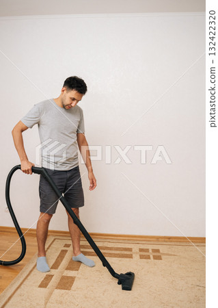 Full length vertical portrait of man vacuuming carpet in living room, focusing on maintaining cleanliness and hygiene, tackling household chores and keeping space tidy. Concept of healthy lifestyle. Full length vertical portrait of man vacuuming carpet in living room, focusing on maintaining cleanliness and hygiene, tackling household chores and keeping space tidy. Concept of healthy lifestyle. 132422320