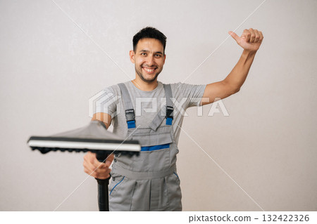Studio portrait of friendly professional cleaning service employee in overall holding vacuum cleaner and showing thumbs up gesture, smiling looking at camera and recommending cleaning company services 132422326