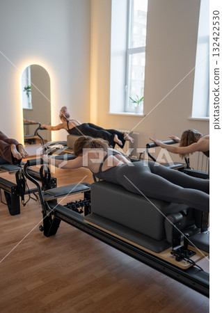 Group of caucasian women exercising with trainer on pilates reformers in a bright studio. Their workout focuses on core strength and flexibility in a modern fitness environment 132422530