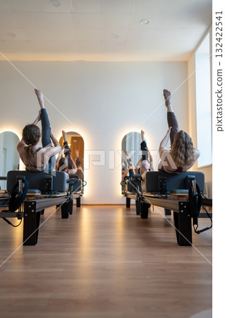 Group of caucasian women exercising with trainer on pilates reformers in a bright studio. Their workout focuses on core strength and flexibility in a modern fitness environment 132422541