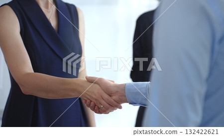 Business professionals shaking hands, sealing a successful partnership agreement, with a colleague applauding in the background against a bright, sunlit window, representing teamwork and achievement Business professionals shaking hands, sealing a successful partnership agreement, with a colleague applauding in the background against a bright, sunlit window, representing teamwork and achievement 132422621
