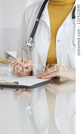 Doctor wearing stethoscope and yellow sweater under white lab coat, making notes on a clipboard in a bright medical office. Medicine, healthcare and science concept Doctor wearing stethoscope and yellow sweater under white lab coat, making notes on a clipboard in a bright medical office. Medicine, healthcare and science concept 132422690
