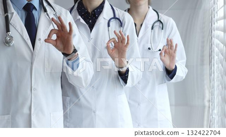 Medical team demonstrating professionalism and successful teamwork, doctors and healthcare workers in lab coats and stethoscopes showing ok sign for quality and well being 132422704
