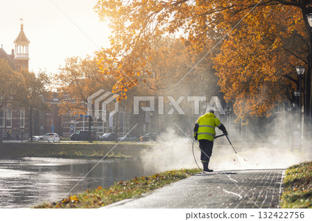 Scenic view municipal worker in yellow uniform cleans sidewalk high pressure washer  fallen leaves after storm rain on sunny fall day at Netherlands city street. Morning scene Dutch urban environment 132422756