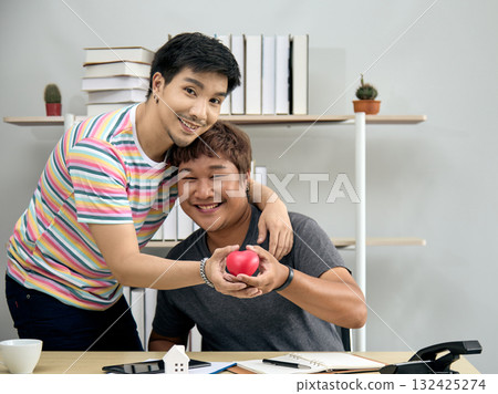Asian gay couple smiling and holding a red heart together while sitting at home office desk. Concept of LGBTQ+ love, relationship support, health awareness, care, and emotional connection Asian gay couple smiling and holding a red heart together while sitting at home office desk. Concept of LGBTQ+ love, relationship support, health awareness, care, and emotional connection 132425274