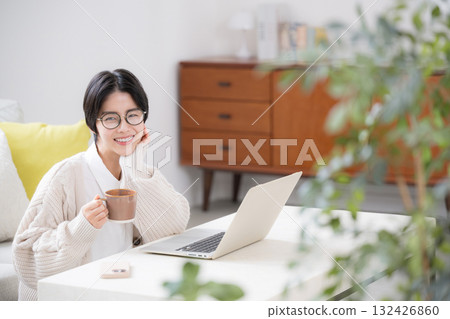 A woman wearing glasses drinking coffee and using a computer in a living room with houseplants, looking at the camera A woman wearing glasses drinking coffee and using a computer in a living room with houseplants, looking at the camera 132426860